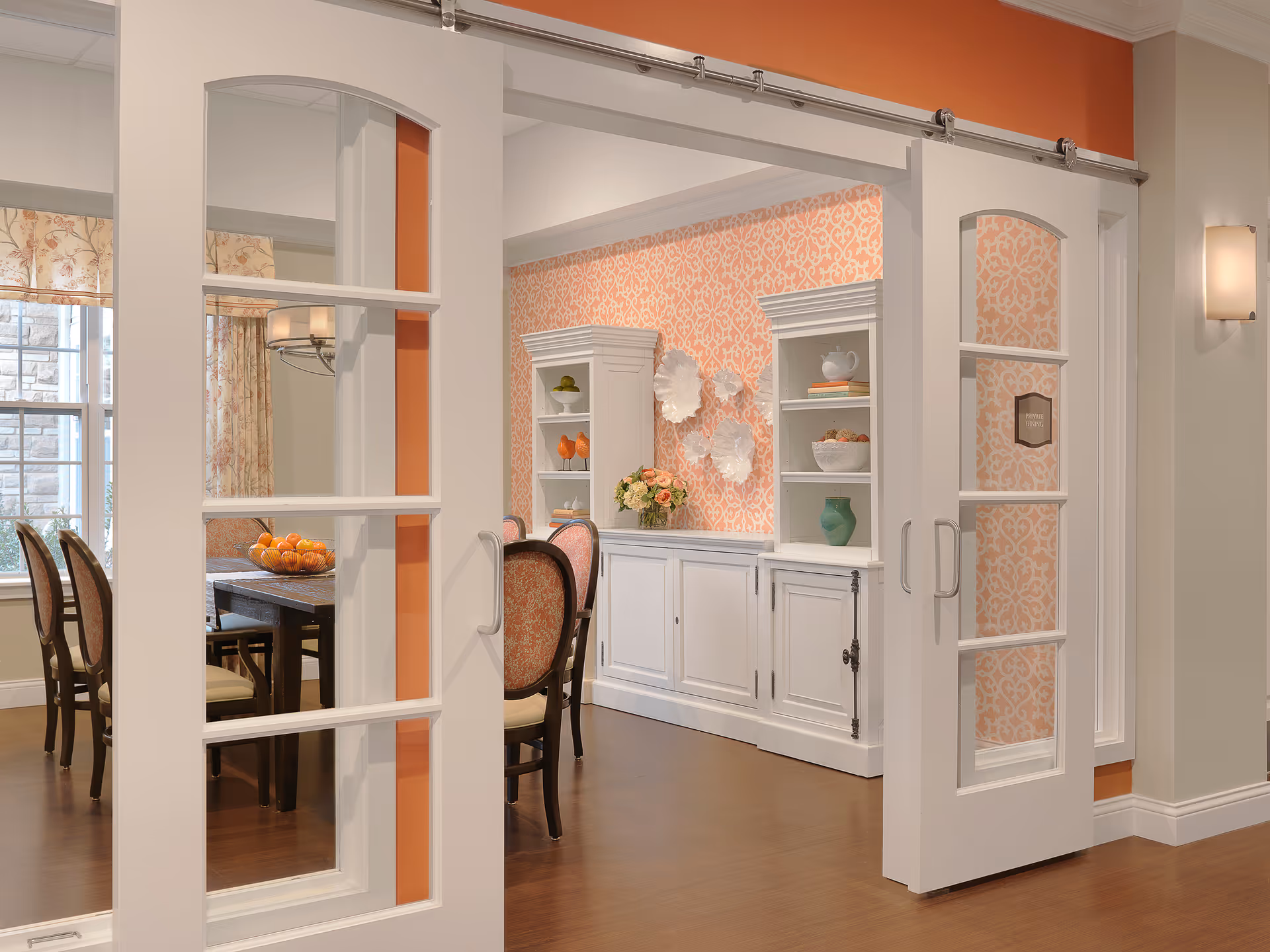 View through white sliding glass-paneled doors into a dining room with a wooden table and chairs. The walls have orange patterned wallpaper, and there are white cabinets with decorative plates and vases. A bowl of oranges is on the table, and a window with floral curtains is visible.