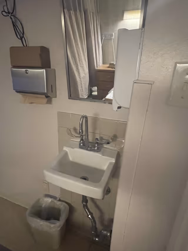 Small wall-mounted sink with exposed plumbing beneath, mirror above, paper towel and soap dispensers on the wall, and a trash can at floor level.