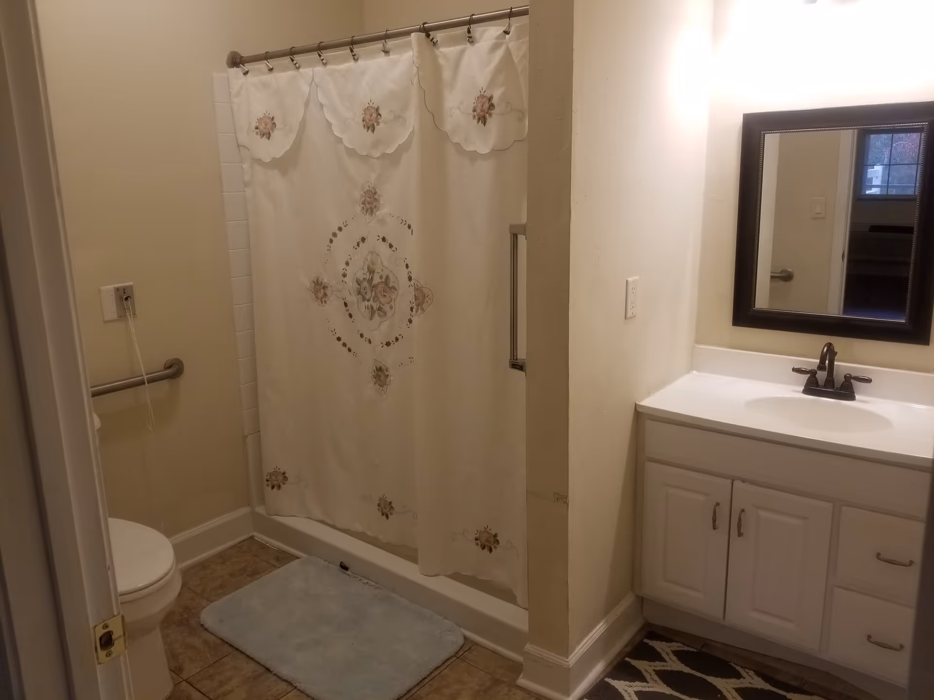 A bathroom with a white toilet, a shower with a floral-patterned curtain, a white vanity with a sink and dark faucet, a mirror above the sink, and beige tiled floor with a light blue bath mat and a black and white rug.