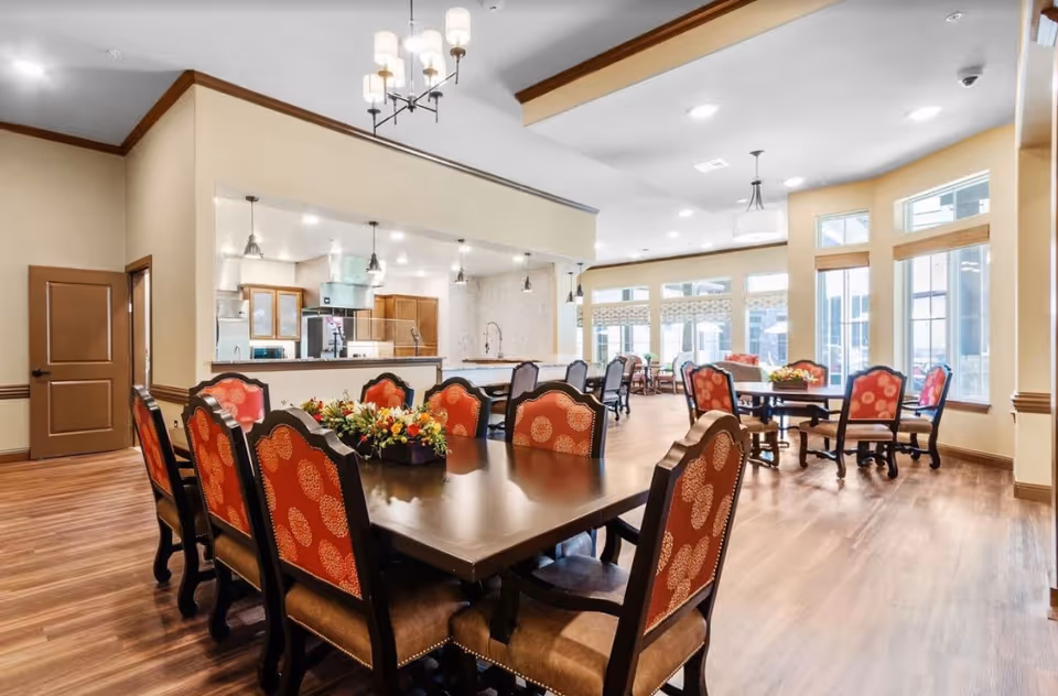 Spacious dining room with multiple wooden tables and red-upholstered chairs, large windows, and an open kitchen area.