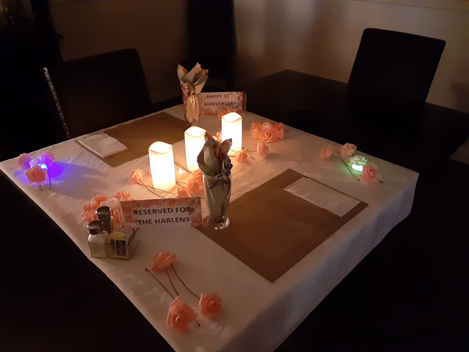 A dining table set for a special occasion with a white tablecloth, brown placemats, folded napkins in glasses, and decorative pink flowers scattered around. Three lit candles are placed in the center, creating a warm ambiance. Two signs on the table read 'HAPPY 50TH ANNIVERSARY' and 'RESERVED FOR THE HARLENS'. Salt and pepper shakers and a small container of sweeteners are also on the table. The room is dimly lit.