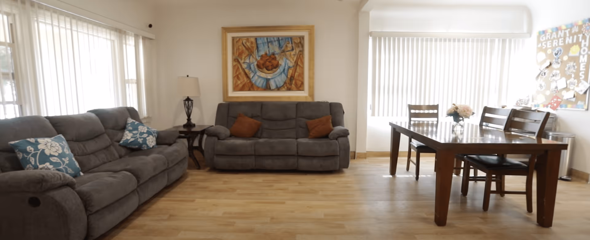 A bright living room area with two gray sofas adorned with decorative pillows, a side table with a lamp, a framed abstract painting on the wall, and a wooden dining table with four chairs near a large window with vertical blinds. A bulletin board on the wall displays the name 'Grant Serenity Homes' with various decorations.