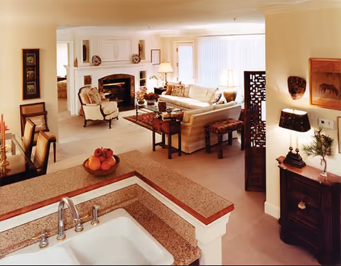A warm and inviting living room area viewed from a kitchen sink countertop. The living room features a white sofa, armchairs, a fireplace with decorative items on the mantel, a coffee table, and lamps providing soft lighting. The space is decorated with framed artwork, plants, and a patterned rug, creating a cozy and comfortable atmosphere.