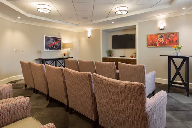 A small theater-style room with two rows of upholstered chairs facing a wall-mounted flat-screen TV. The room has soft lighting with ceiling fixtures and wall sconces. There are framed movie posters on the walls and a console table with a lamp and decorative items.