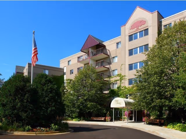 Exterior view of a multi-story senior living facility building with balconies, surrounded by trees and landscaping. An American flag is displayed on a flagpole near the entrance, which has a covered awning. The sky is clear and blue.