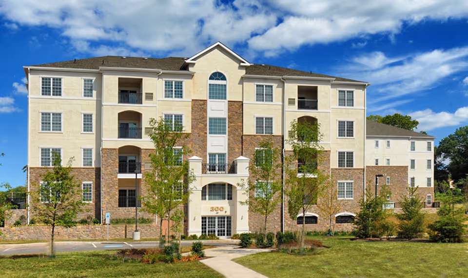 Front exterior view of a multi-story residential building with stone and stucco facade, balconies, and landscaped lawn under a blue sky.