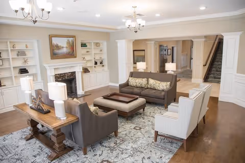 A spacious and well-lit living room with a fireplace, built-in white shelves, and a large painting above the mantel. The room features a seating arrangement with two brown sofas, two white armchairs, a wooden coffee table, and two table lamps on a wooden console table. The floor is hardwood with a patterned area rug, and there are chandeliers hanging from the ceiling.