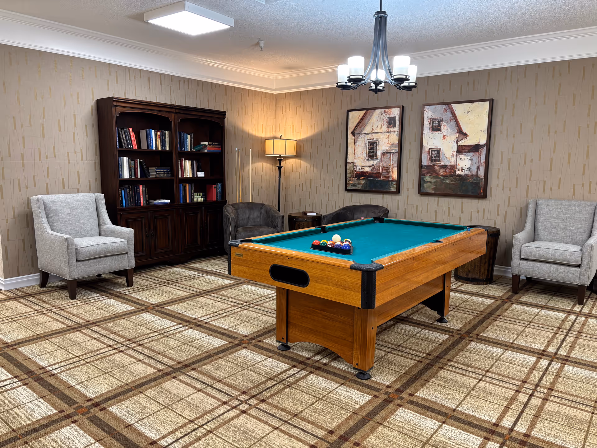 A cozy room with a pool table in the center, surrounded by two gray armchairs, two dark brown chairs, a wooden bookshelf filled with books, a floor lamp, and two framed paintings of houses on the wall. The room has patterned carpet flooring and a modern chandelier hanging from the ceiling.