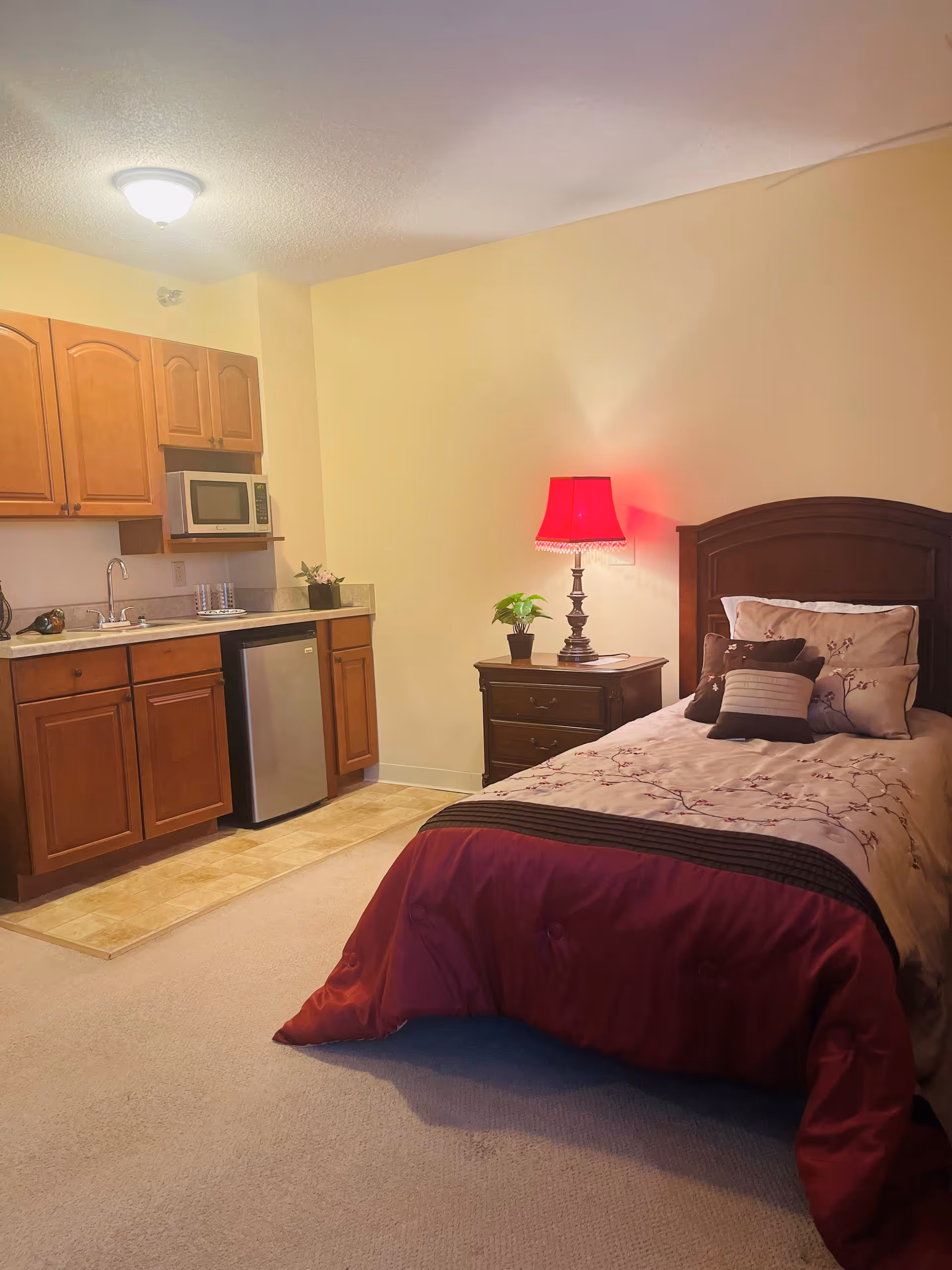 A cozy room featuring a single bed with floral and striped pillows and a red comforter. Next to the bed is a wooden nightstand with a potted plant and a lamp with a red lampshade. The room also includes a small kitchenette area with wooden cabinets, a sink, a microwave, and a mini refrigerator. The walls are painted light yellow and the floor is carpeted except for the kitchenette area which has tile flooring.