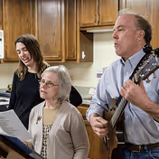 Three people in a kitchen setting, with a man playing a guitar and singing, and two women standing nearby, one holding a sheet of paper and singing along.
