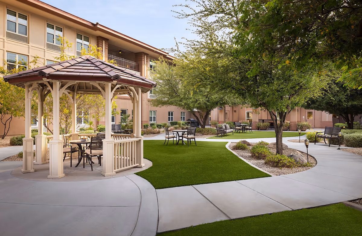 Outdoor courtyard area at Belmont Village Senior Living Scottsdale featuring a gazebo with seating, multiple tables and chairs, green artificial turf, trees, and a surrounding beige building.