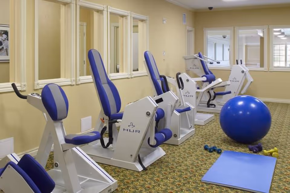 A small exercise room with several pieces of seated fitness equipment with blue and gray padding, a large blue exercise ball, a blue exercise mat, and several small dumbbells on a patterned carpet floor. The walls are light yellow with white trim and multiple windows.