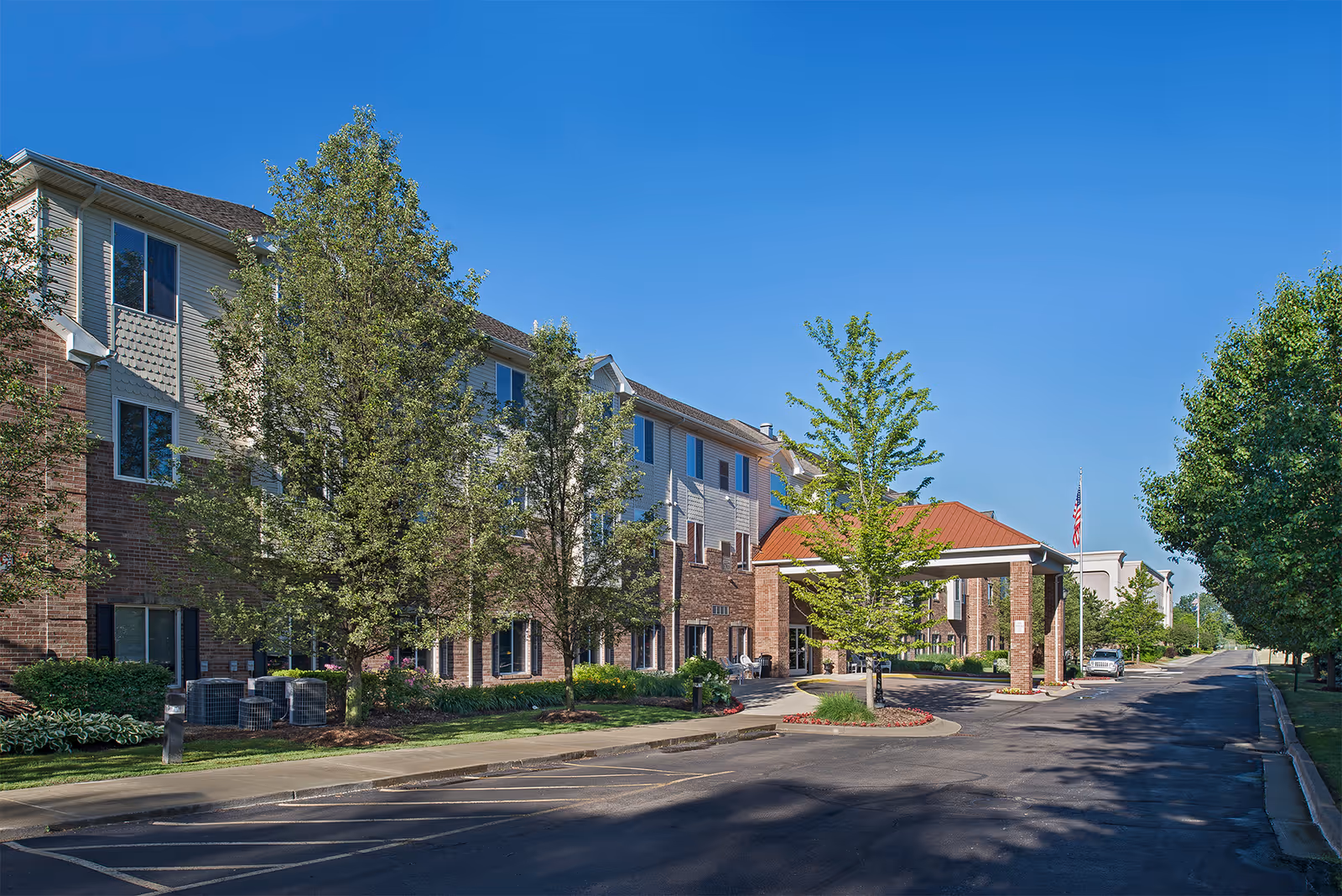 Exterior view of a multi-story senior living facility building with a covered entrance, surrounded by trees and landscaping under a clear blue sky.