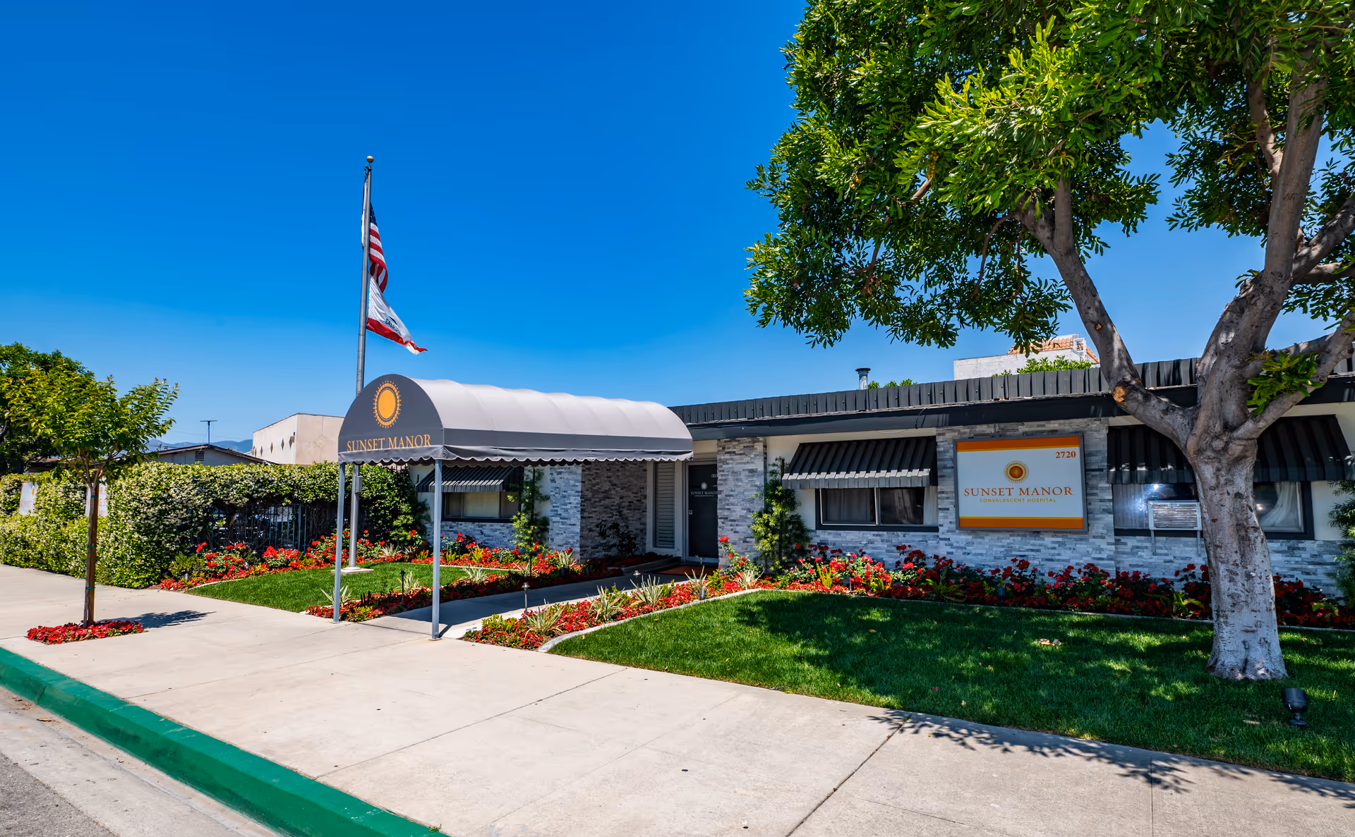 Exterior view of Sunset Manor senior living facility on a sunny day with a clear blue sky. The building has a white and gray brick facade with a covered entrance canopy displaying the Sunset Manor logo. There are well-maintained green lawns, flower beds with red flowers, a tree, and two flags on a flagpole near the entrance.