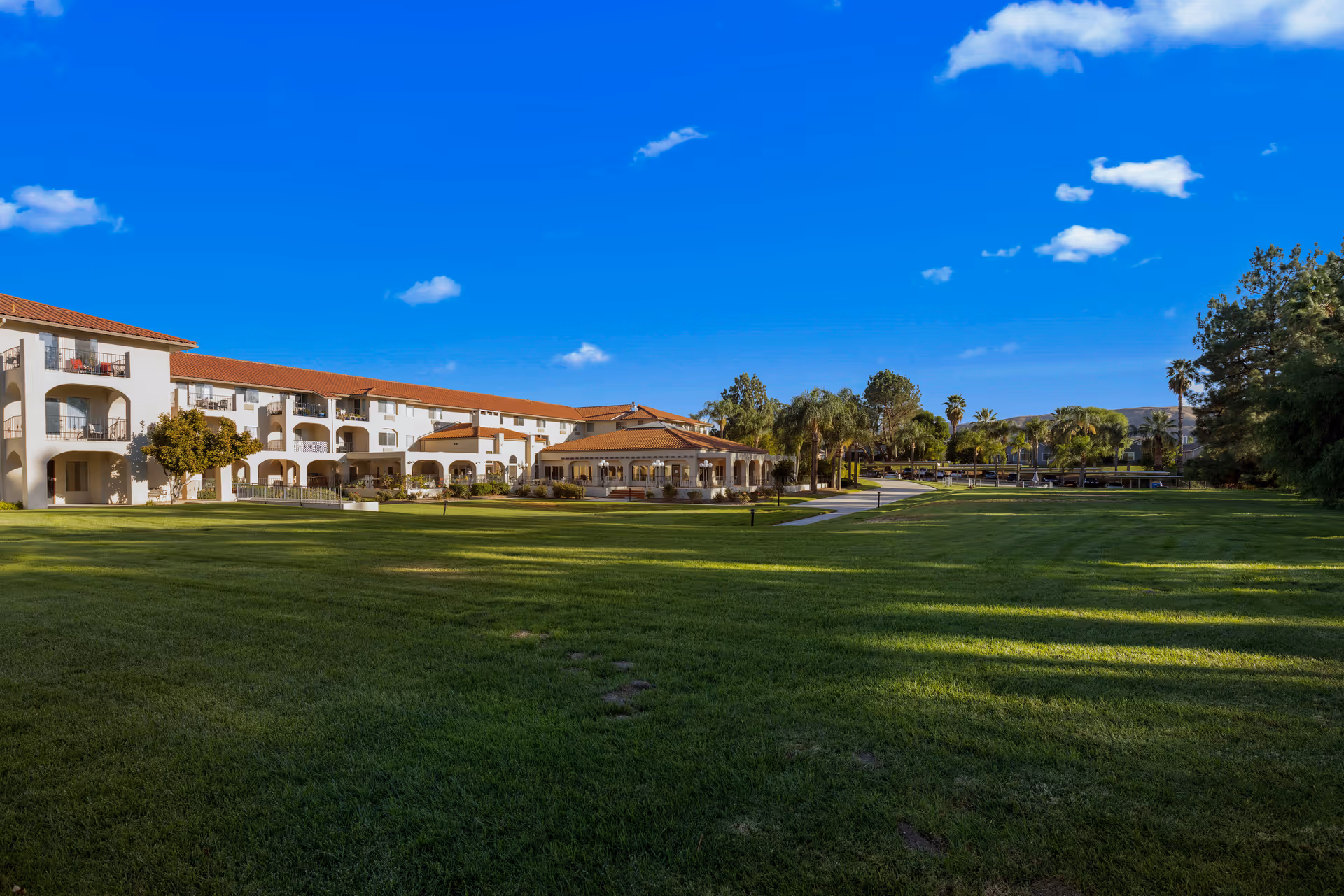 A large green lawn with a paved walkway leading to a two-story building with white walls and red-tiled roofs under a bright blue sky with a few scattered clouds. The building has balconies and arches, surrounded by trees and landscaping.