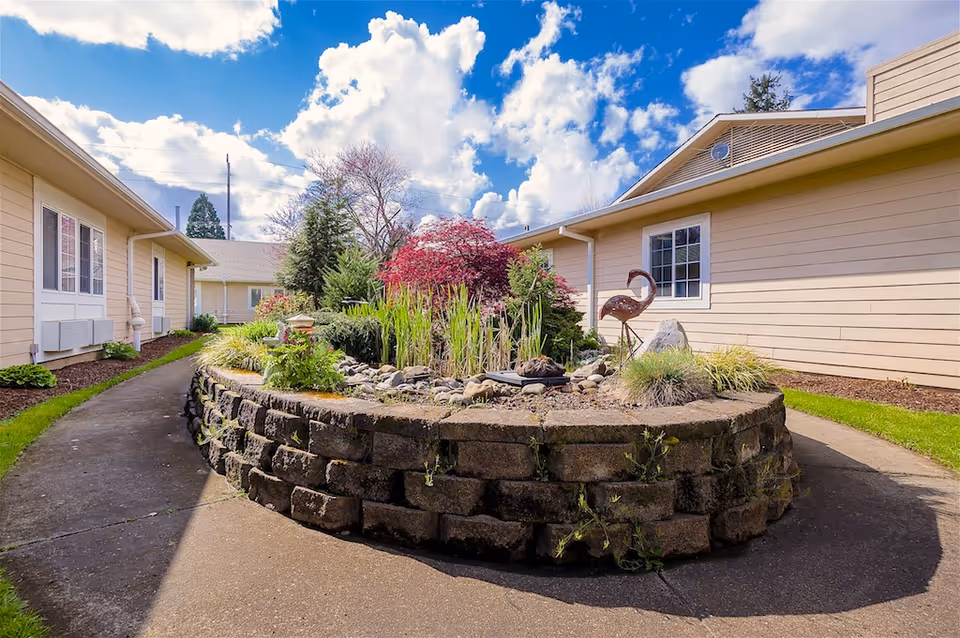 Sunlit courtyard between single-story senior living buildings with a raised stone planter filled with plants and a metal flamingo statue under a blue sky.
