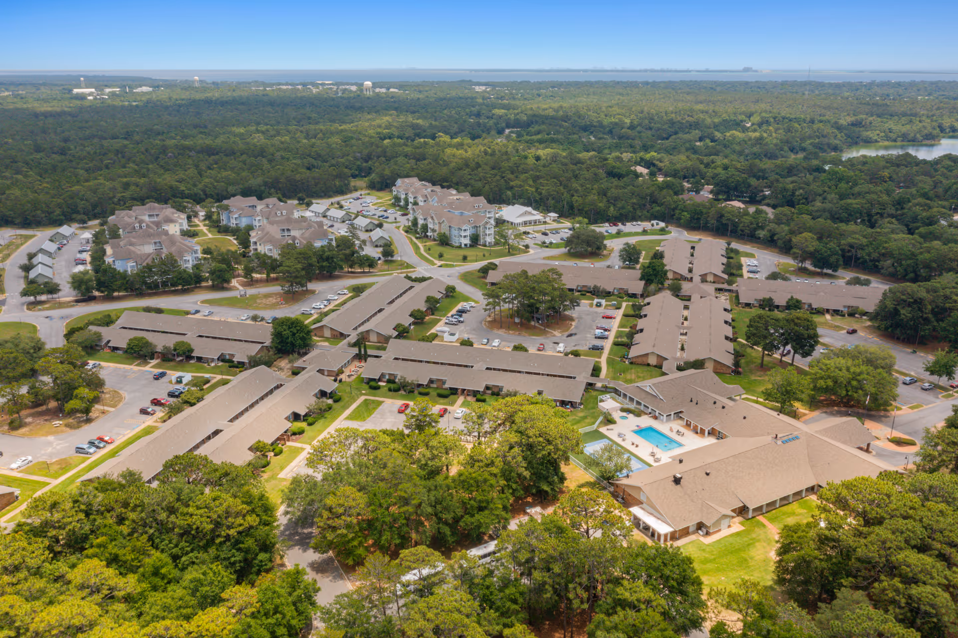 Aerial view of the Air Force Enlisted Village senior living facility showing multiple residential buildings with brown roofs, parking lots with cars, a swimming pool, and surrounding green trees and forested area extending to the horizon.