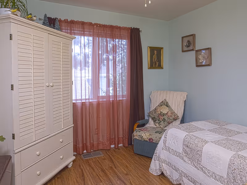 A cozy bedroom in a senior living facility featuring a bed with a patterned quilt, a cushioned armchair with a floral pillow and a white throw, a white wooden wardrobe, and a window with sheer pink curtains allowing natural light to enter. The walls are light blue and decorated with framed artwork.