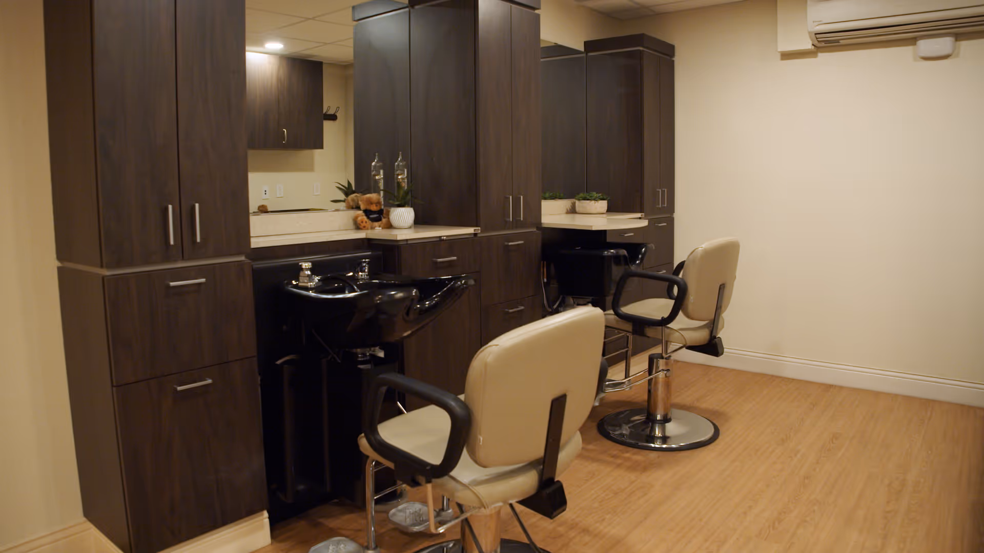 Interior view of a salon area in a senior living facility featuring two beige salon chairs with black armrests facing dark wood cabinetry and black wash basins. The room has light-colored walls, a wooden floor, and an air conditioning unit mounted on the wall.