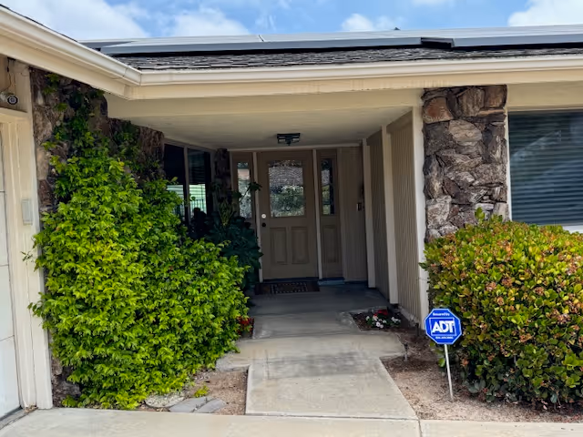 Covered front entrance of a single-story building with a centered door, stone columns, shrubs, and an ADT security sign.