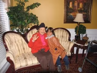 An elderly woman and a young girl sitting together on a beige upholstered sofa in a warmly decorated living room. The elderly woman is wearing a red sweater and a black hat, smiling and waving at the camera. The young girl is leaning close to her, wearing a striped jacket. Behind them is a large green plant, a side table with a lamp, and a framed painting on a yellow wall.