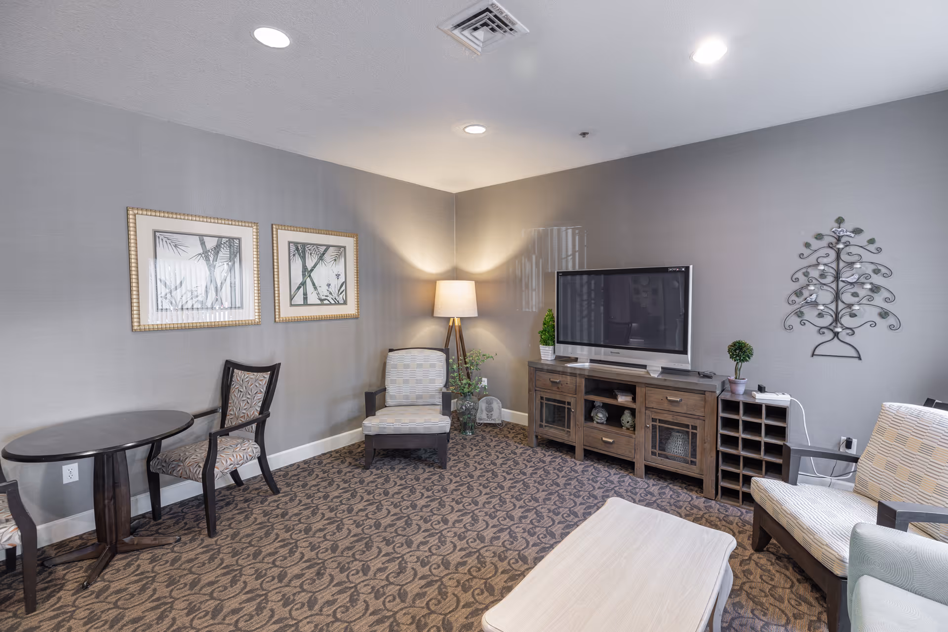 Cozy common area with upholstered chairs, a coffee table, a TV on a wooden console, a floor lamp, wall art, and patterned carpet.