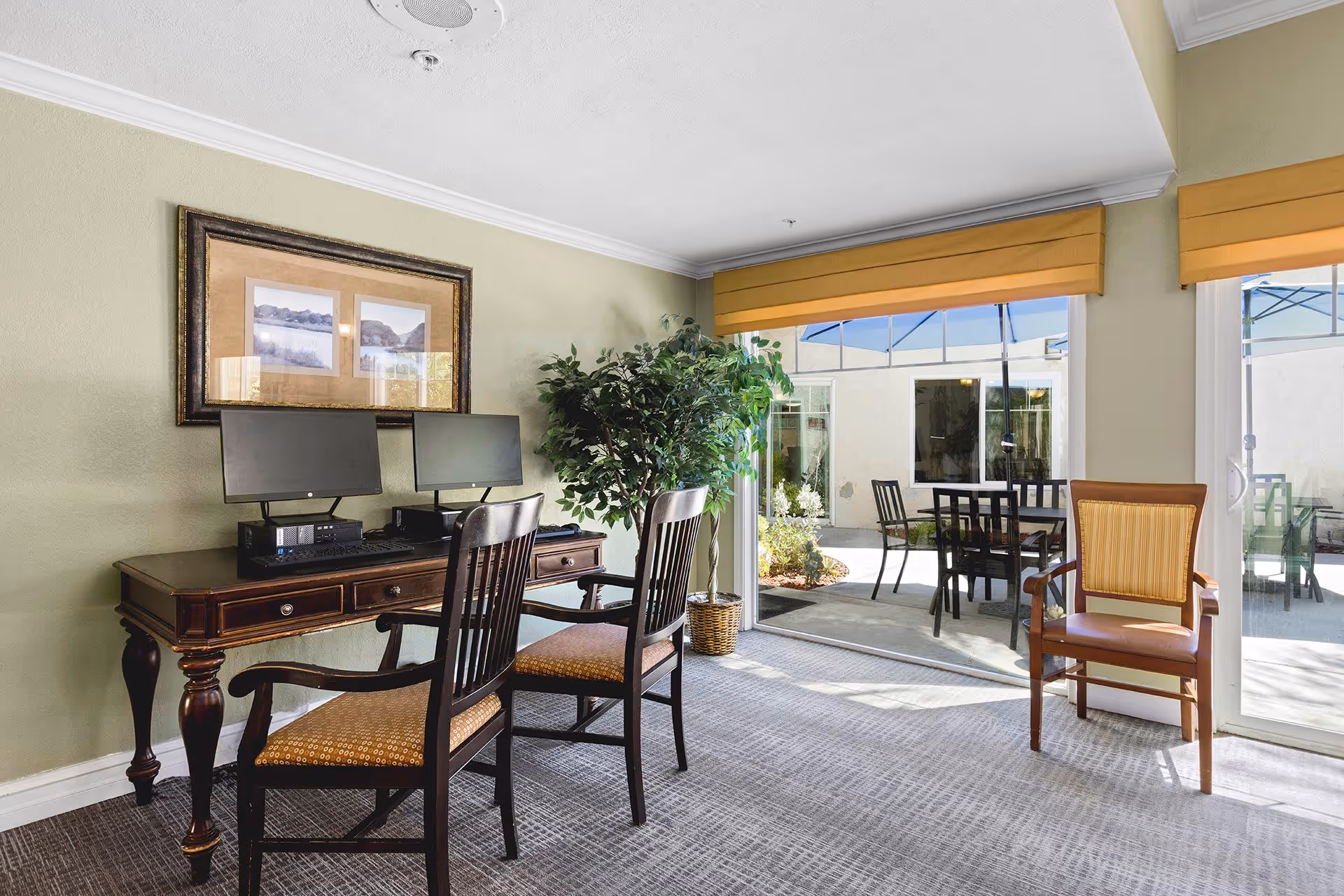 A bright room with two computer workstations on a wooden desk, each with a monitor and keyboard. Two wooden chairs with cushioned seats are placed in front of the desk. A large potted plant is next to the desk. The room has light green walls, a framed picture above the desk, and large sliding glass doors with yellow valances that open to an outdoor patio area with tables, chairs, and umbrellas.