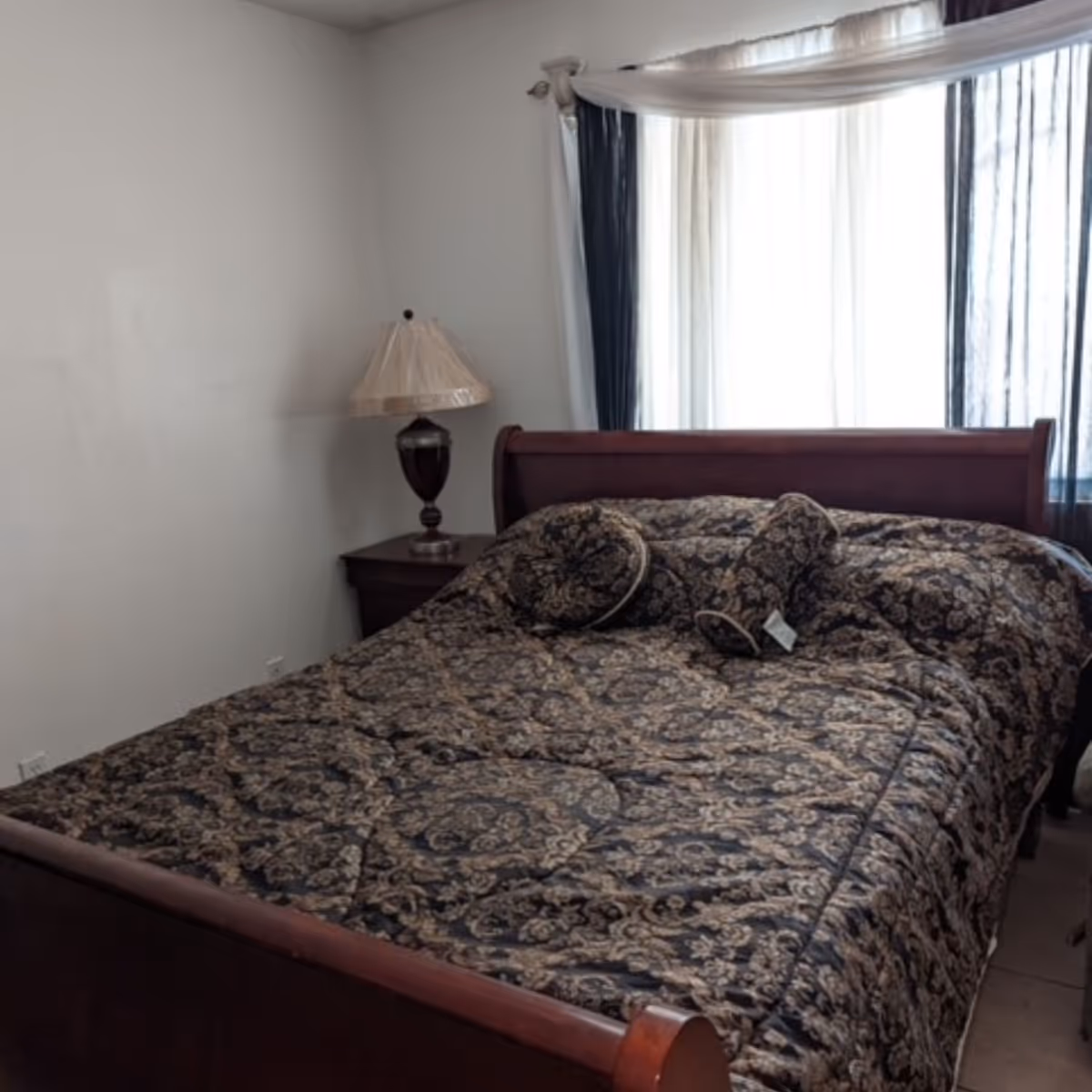 Neatly made bed with patterned bedding, nightstand, lamp, and draped window in bedroom.