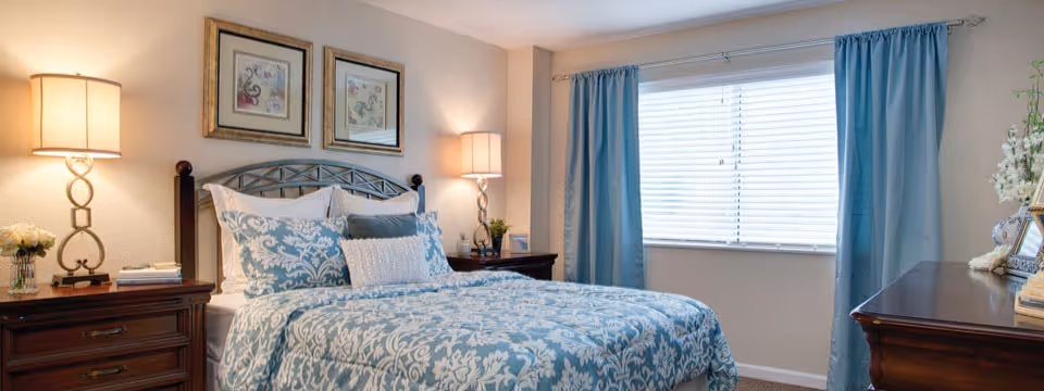 Bright bedroom with a bed dressed in blue-and-white patterned bedding, wooden nightstands with lamps, framed art above the headboard, and a window with blue curtains.