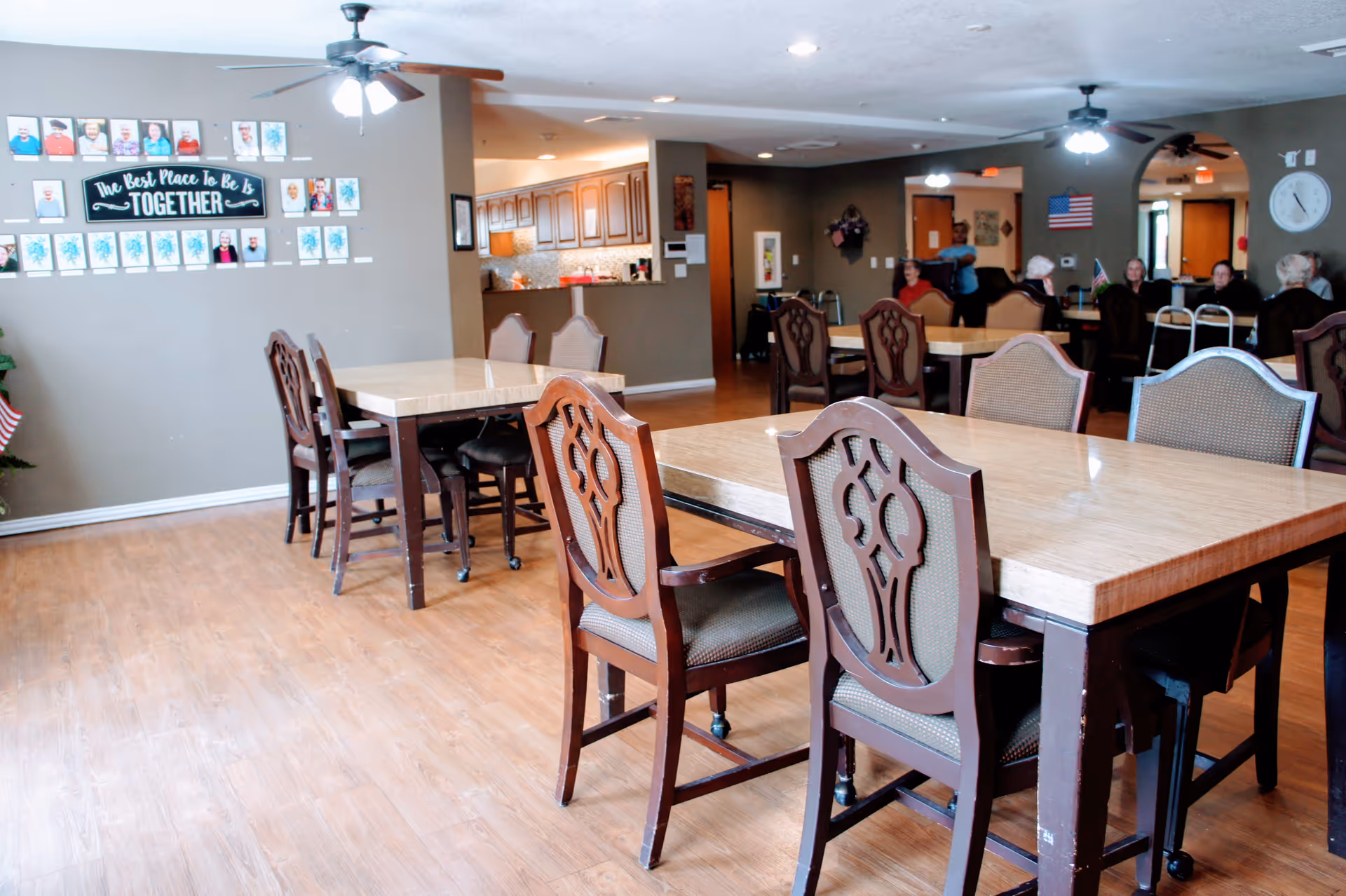 Interior view of a senior living facility dining area with several wooden tables and chairs. In the background, a group of elderly people are seated and a caregiver is assisting one of them. The walls are decorated with framed photos and a sign that reads 'The Best Place To Be Is TOGETHER'. Ceiling fans with lights hang from the ceiling.