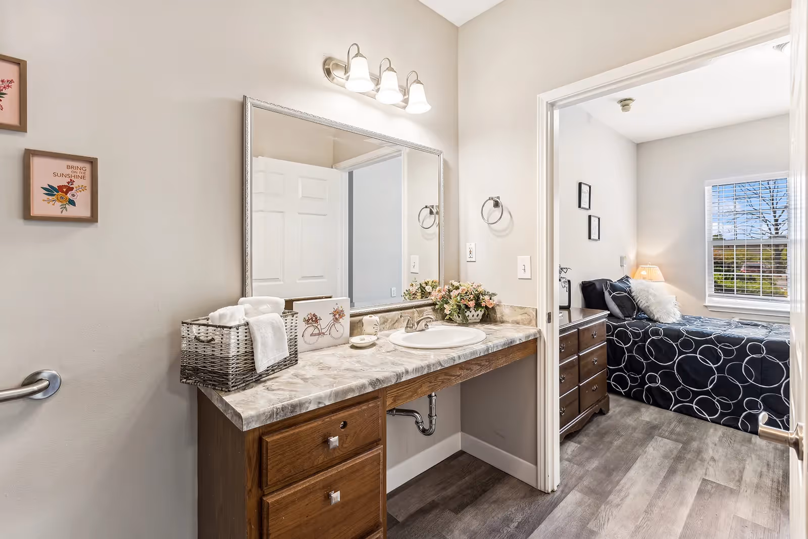 A bathroom vanity area with a large mirror, marble countertop, and wooden drawers. A basket with towels and decorative items are on the counter. Through an open door, a bedroom with a bed covered in a black and white circular patterned bedspread, a wooden dresser, and a window with blinds is visible.