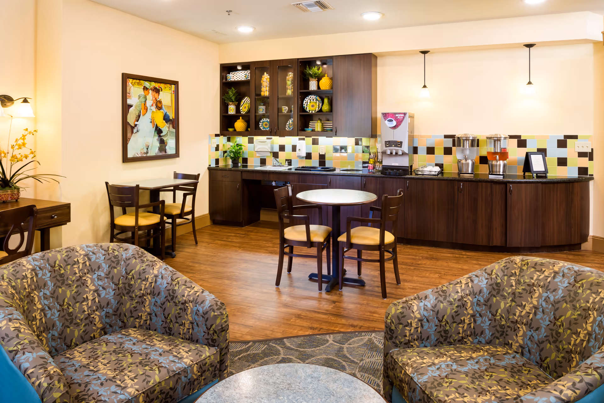 A cozy common area with patterned armchairs and a small round table in the foreground. In the background, there is a kitchenette with dark wood cabinets, a colorful tiled backsplash, a coffee machine, and beverage dispensers. There are also two small dining tables with chairs, a framed painting on the wall, and warm lighting fixtures.