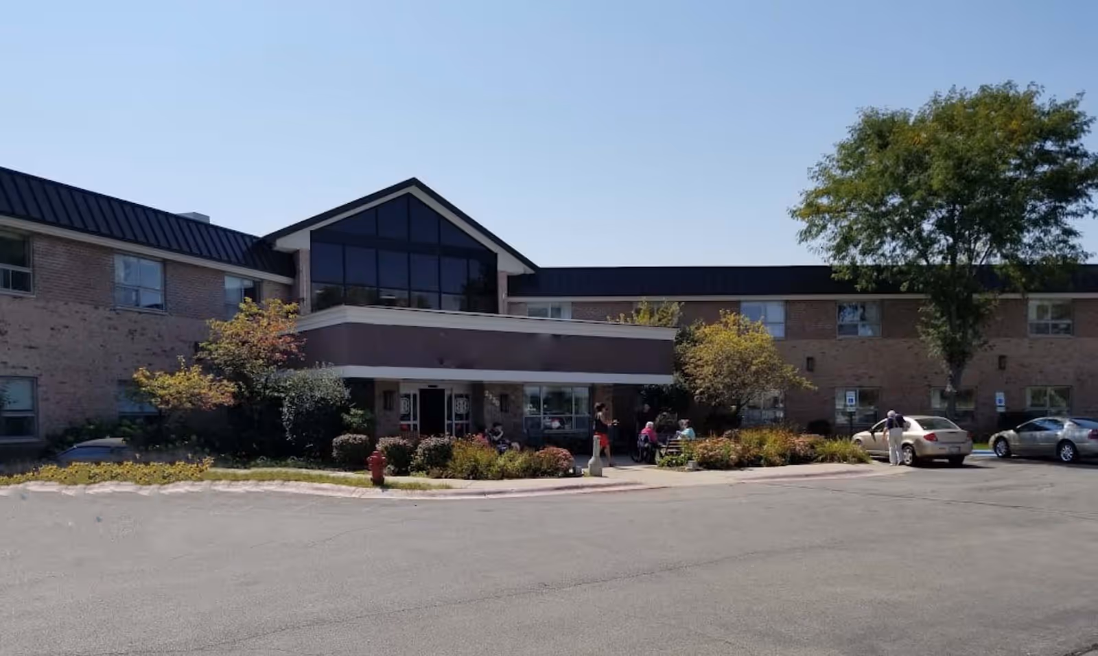 Front exterior of a two-story brick senior living building with a covered entrance, landscaping, parked cars and people gathered outside.