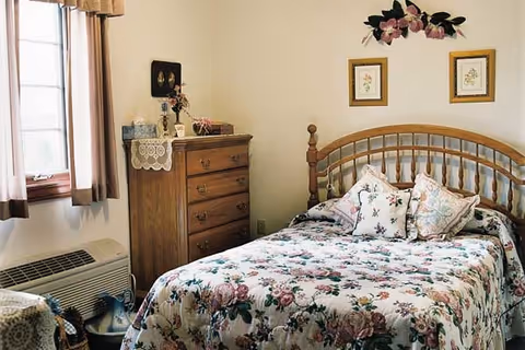 A cozy bedroom with a wooden bed frame and a floral bedspread. The bed has several decorative pillows. There is a wooden chest of drawers with a lace doily and decorative items on top. Two framed floral pictures hang on the wall above the bed, and a floral wall decoration is above them. A window with curtains lets in natural light, and an air conditioning unit is below the window.