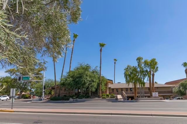 Exterior view of Heritage Court Post Acute Of Scottsdale facility with a clear blue sky, palm trees, and other greenery surrounding the building. The building has a low, wide structure with a brown roof and is situated next to a street with traffic signals and road signs.