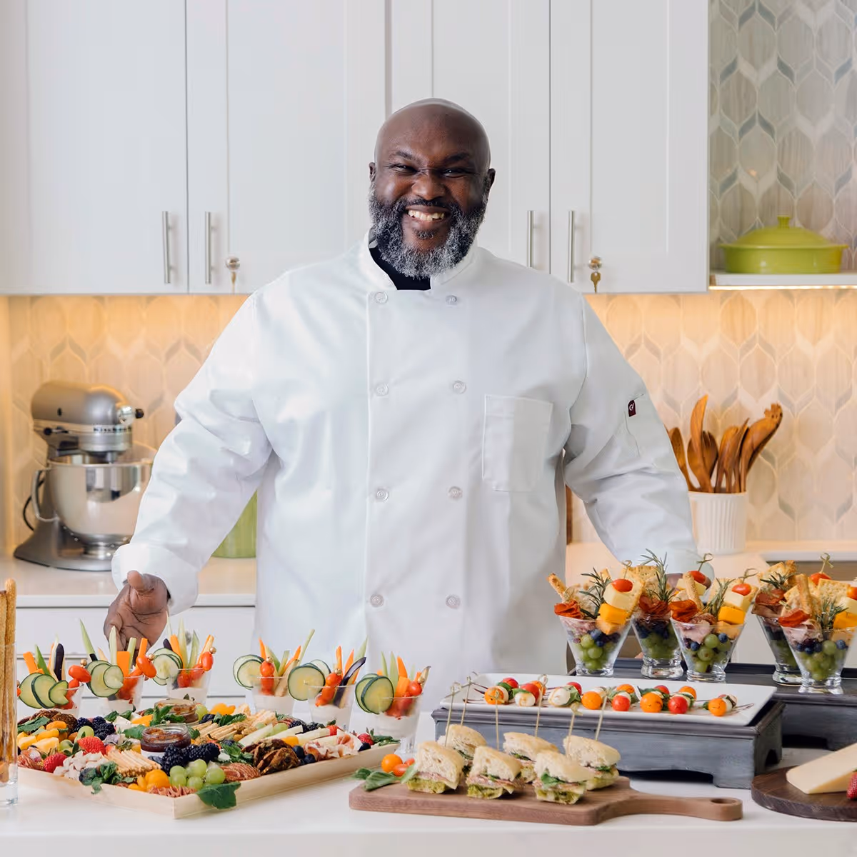 A smiling chef in a white coat stands behind a kitchen counter filled with an assortment of colorful appetizers including vegetable cups, sandwiches, fruit and cheese skewers, and parfait glasses with fruit and cheese. The background shows white kitchen cabinets, a mixer, and wooden utensils.