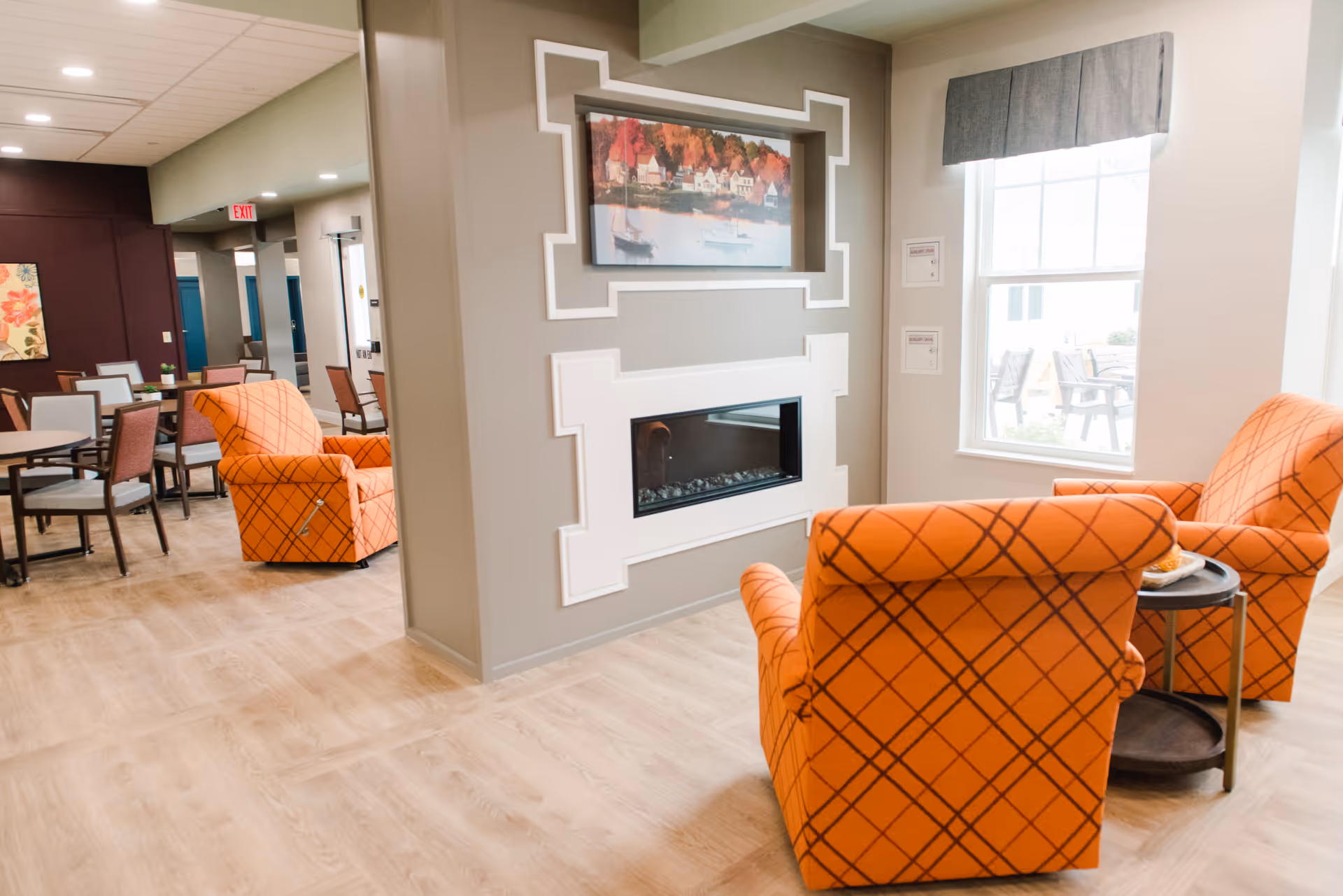 Bright senior living common area with orange patterned armchairs facing a built-in fireplace and dining tables visible in the background.