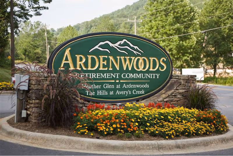A large green and gold sign for Ardenwoods Retirement Community, surrounded by colorful flowers and landscaping, with trees and a road in the background.