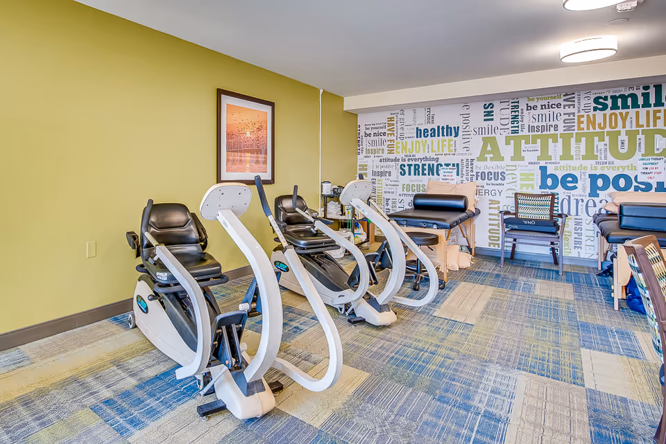 A fitness room with three recumbent exercise bikes lined up on a patterned carpet. The walls are painted light green and white, with one wall featuring a large motivational word collage including words like 'attitude', 'smile', and 'be positive'. There are also two padded treatment tables and a chair with a cushion against the wall.