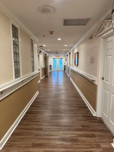 A long, well-lit hallway with wood flooring and beige walls featuring white handrails on both sides. The ceiling has recessed lighting, vents, and a smoke detector. At the end of the hallway, there is a glass door with a red exit sign above it.