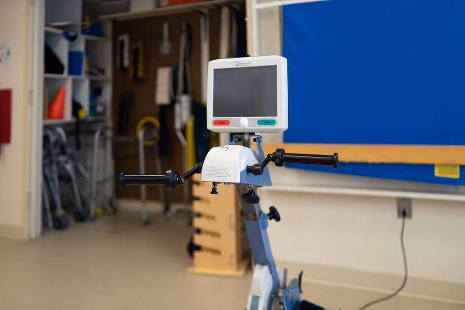 A stationary upper-body rehabilitation exercise machine with a monitor and handlebars in a therapy room with walkers and equipment shelves behind it.