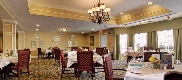 A spacious dining room with multiple tables covered in white tablecloths, set with glasses, cups, and floral centerpieces. The room features patterned carpet, large windows with green curtains allowing natural light, and a decorative chandelier hanging from the ceiling. Framed artwork and a round mirror adorn the beige walls.