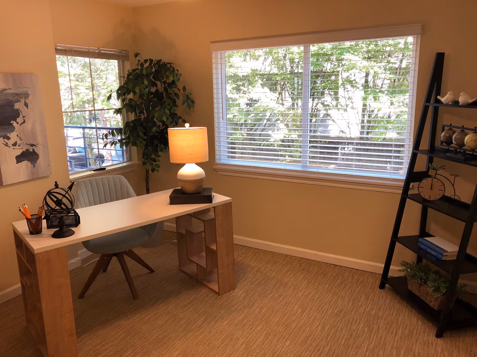 A bright office space with a modern desk and a comfortable chair. On the desk, there is a lamp, a pencil holder with pencils, and a decorative globe. The room has two large windows with white blinds, allowing natural light to fill the space. A tall green plant is placed near one window, and a black ladder-style shelf with books, decorative globes, a clock, and small plants is positioned against the wall.
