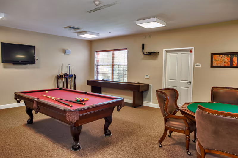 A recreational room with a red felt pool table in the center, pool cues resting on the table and in a rack against the wall. There is a shuffleboard table under a window with blinds, a wall-mounted TV, and a round table with four brown leather chairs. The walls are beige and the floor is carpeted.