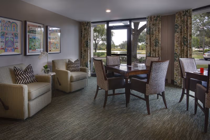 A cozy interior room with two beige armchairs featuring patterned pillows on the left, a wooden table with four upholstered chairs in the center, and large windows with floral curtains showing trees and a parking lot outside.