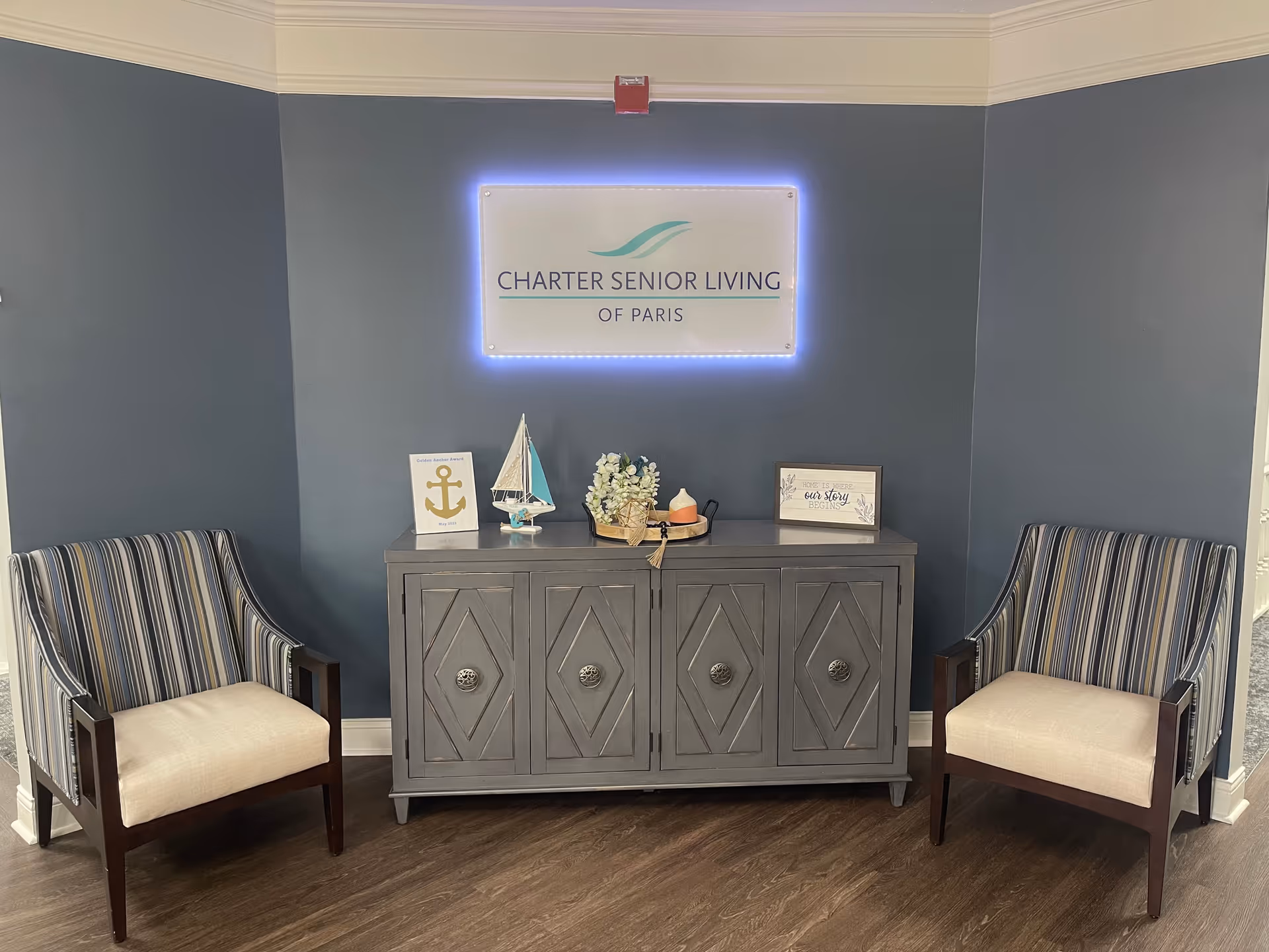 A cozy corner in Charter Senior Living of Paris featuring two striped armchairs with beige cushions on either side of a gray wooden cabinet. On top of the cabinet are decorative items including a small sailboat model, a basket with flowers, and framed signs. Above the cabinet is a lit sign displaying the facility's name.