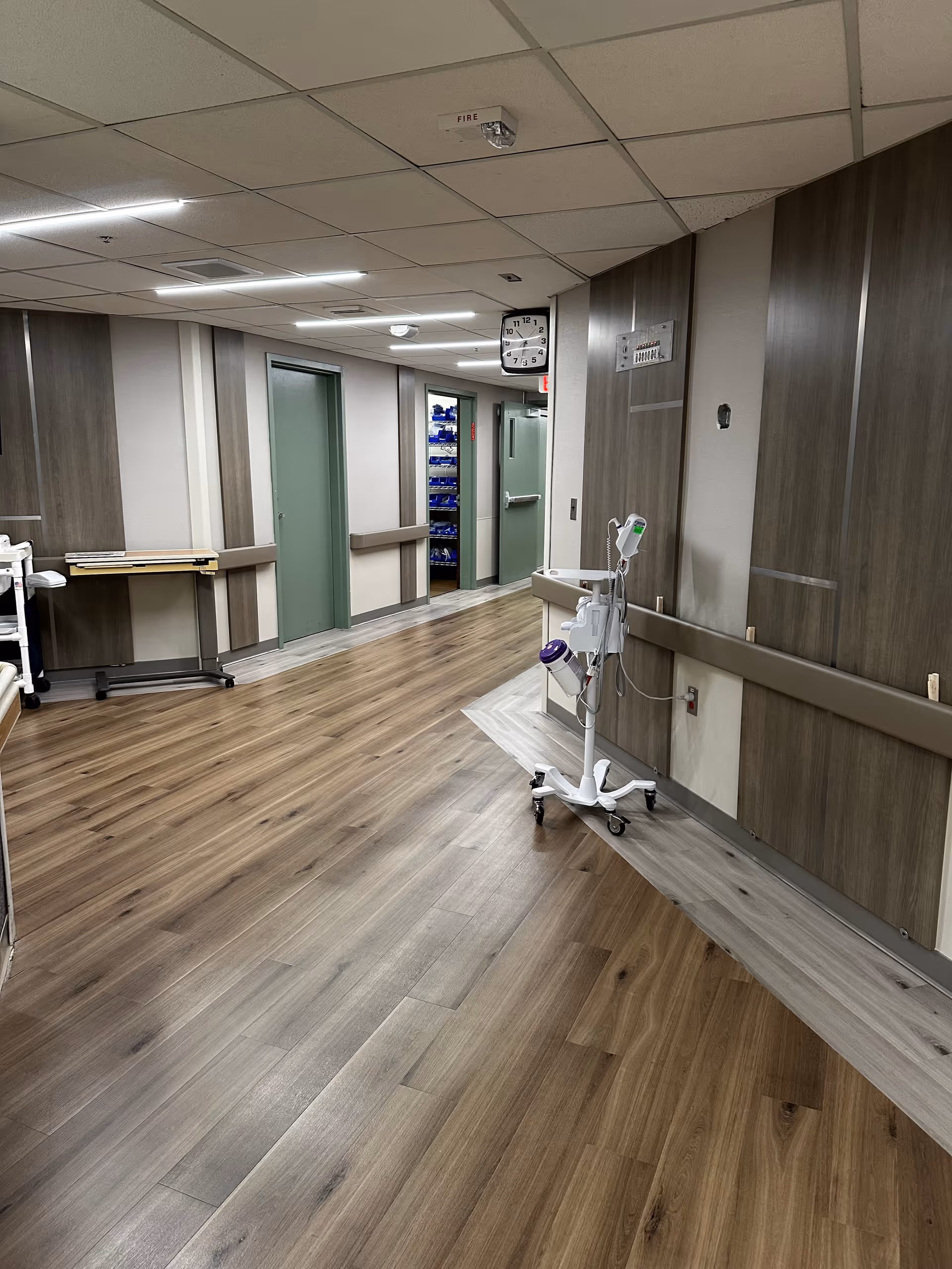 A clean, well-lit hallway in a healthcare facility with wood-patterned flooring and light-colored walls. There are several closed green doors along the hallway, a clock mounted on the wall, and a medical equipment stand on wheels positioned near the right wall.