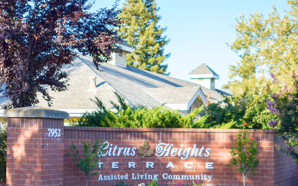 Brick entrance sign for 'Citrus Heights Terrace Assisted Living Community' with shrubs, trees, and the facility roof visible behind it.