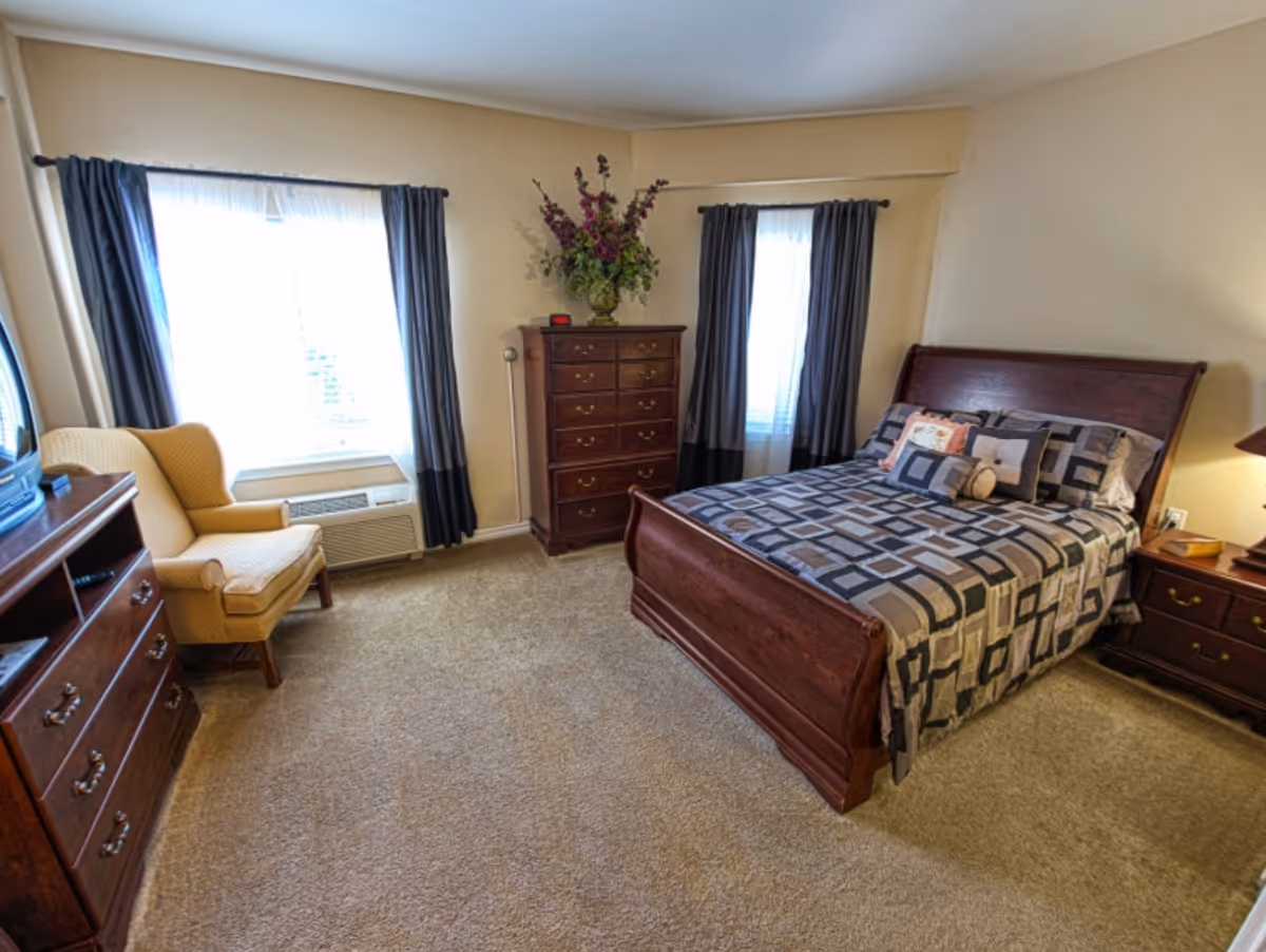 A cozy bedroom with a wooden bed frame and a patterned bedspread, two windows with dark curtains, a wooden dresser with a vase of flowers on top, a yellow upholstered armchair, a wooden chest of drawers with a TV on top, and beige carpeted floor.