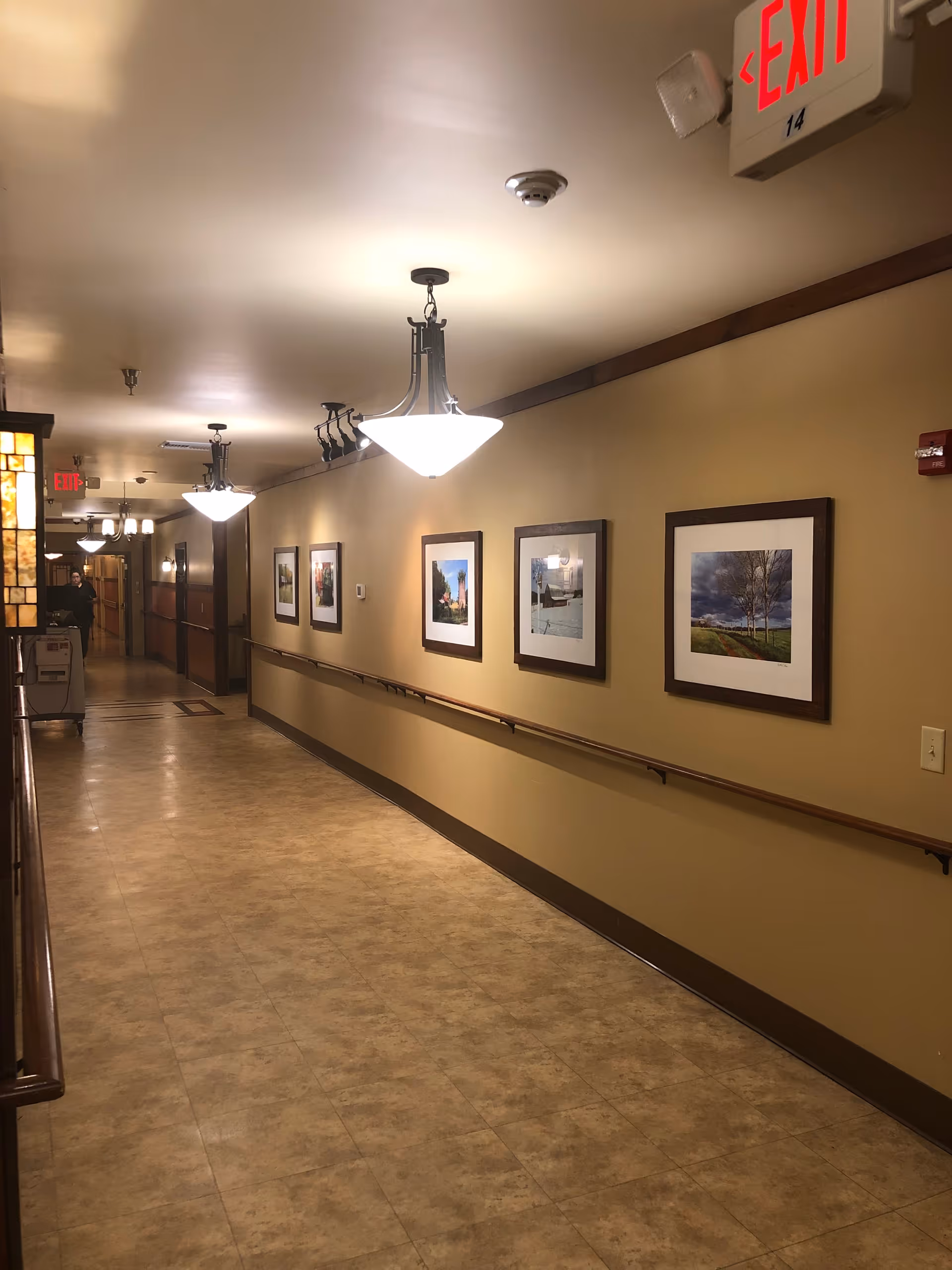 Well-lit interior hallway with framed artwork, handrails along the walls, and illuminated exit signs.